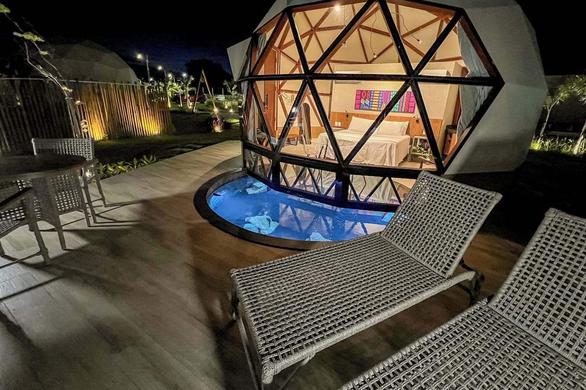 Una estructura de domo geodésico con piscina, sillones de descanso y una cama a la vista en su interior.
