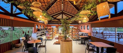 Un restaurante con techos altos, lámparas colgantes de mimbre y una planta en maceta en el centro. Mesas y sillas dispuestas para comer.