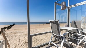 Blick auf einen Strand aus einer gläsernen Veranda mit Tisch und Stühlen, einer Hängelampe und einer Holzterrasse, die zum Strand führt.