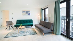 Ein modernes Wohnzimmer mit einem grünen Sofa, einem grauen Sofa, einem Holztisch und einem kleinen Sessel. Ein großes Fenster bietet Blick auf Gebäude und einen Balkon.
