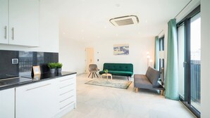 Eine moderne Küche mit weißen Schränken, einer schwarzen Arbeitsplatte und einer kleinen Kochinsel mit Pflanzen. Im Wohnbereich befindet sich ein grünes Sofa und ein Sessel, mit einem großen Fenster, das einen Ausblick bietet.