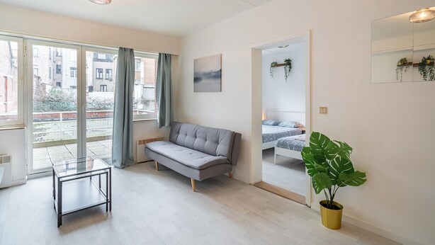 Ett modernt vardagsrum med en soffa, ett soffbord i glas, ett stort fönster, en krukväxt och en säng i det intilliggande rummet.