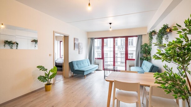 Een moderne woonkamer met een bank, een eettafel, stoelen, een potplant en een groot raam met gordijnen.