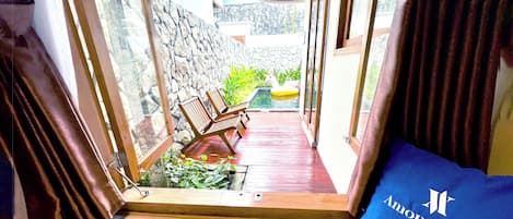 A corridor with wooden flooring, a stone wall, and a small pool with a white inflatable unicorn. There are wooden chairs and a blue pillow with 'Among Villas' text.