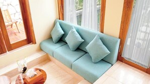 Un canapé bleu clair agrémenté de quatre coussins, une table d’appoint en bois sur laquelle est posé un vase de fleurs blanches, et une vue par une fenêtre ornée de rideaux légers.
