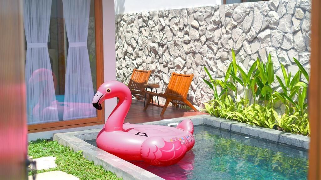 A swimming pool with a pink flamingo float, outdoor chairs, a stone wall, and green plants.
