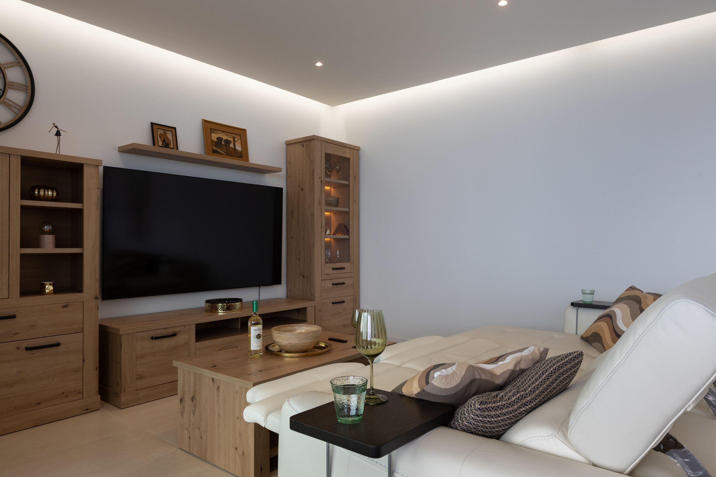 Ein modernes Wohnzimmer mit einem Flachbildfernseher, Holzschränken, einem Glastisch und einer Couch.