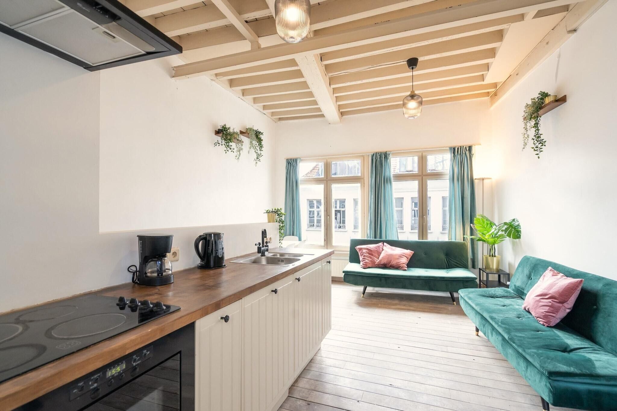 A modern kitchen with a wooden countertop, a built-in stove, and a sink. There is a green sofa with pink pillows in the living area, and a window with curtains.