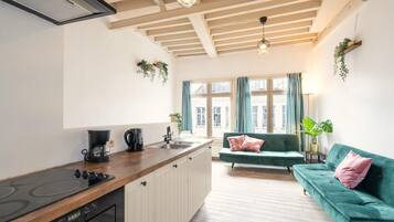 A modern kitchen with a wooden countertop, a built-in stove, and a sink. There is a green sofa with pink pillows in the living area, and a window with curtains.