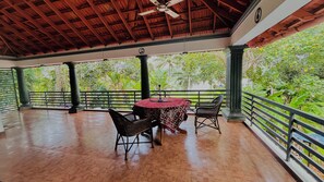 A covered outdoor area with a wooden floor, a table with chairs, and a view of greenery. - Harbour villa (Thiruvananthapuram)