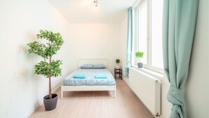 Une chambre à coucher avec un lit, une plante en pot, une petite table sur laquelle est posée une plante, et une fenêtre avec des rideaux.