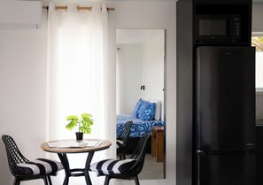 Ein kompakter Raum mit Esstisch, zwei Stühlen, einem schwarzen Kühlschrank und einem Bett mit blauer Bettwäsche.