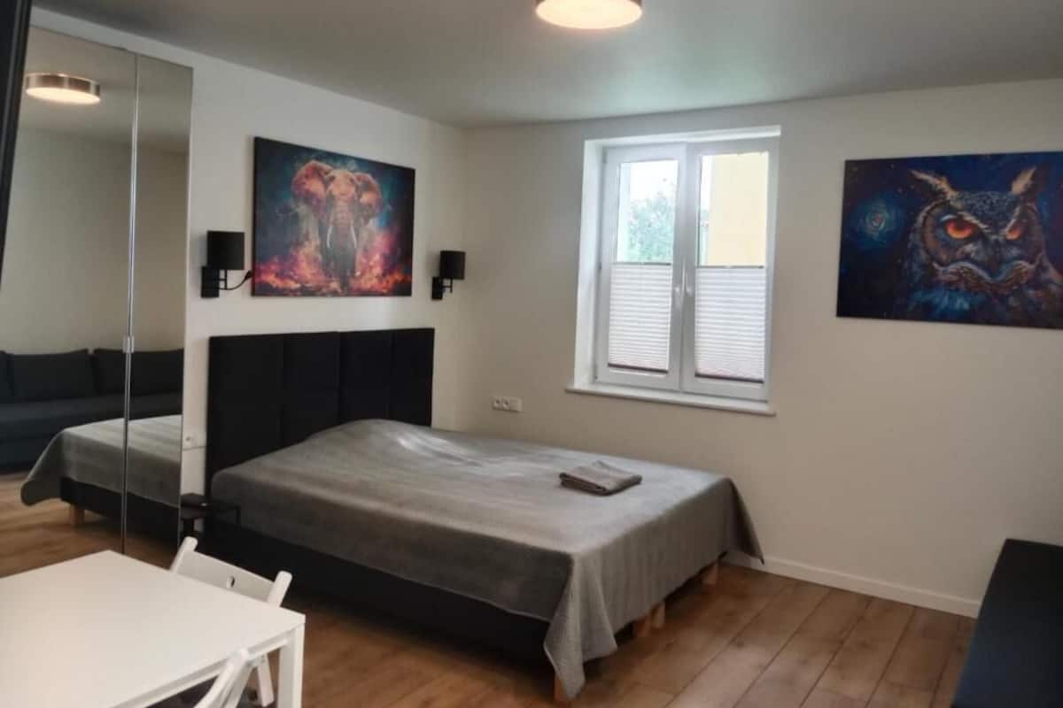 A bedroom with a bed, a desk, a chair, a window, and two paintings on the walls.