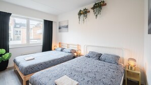 Quarto de hotel com duas camas, janela com cortinas, radiador, planta suspensa na parede e mesa de cabeceira com abajur.