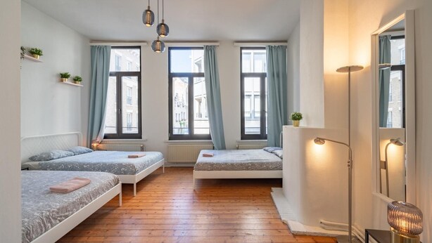 Een kamer met drie bedden, houten vloeren, een staande lamp en een groot raam met gordijnen.