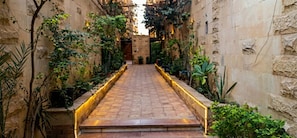 Un sendero estrecho bordeado por muros de piedra y plantas en macetas, que lleva a la entrada de un edificio.