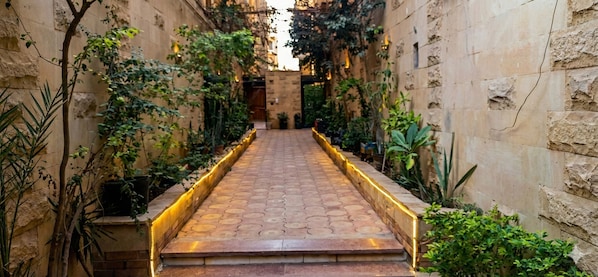A narrow pathway flanked by stone walls and potted plants, leading to a building entrance.