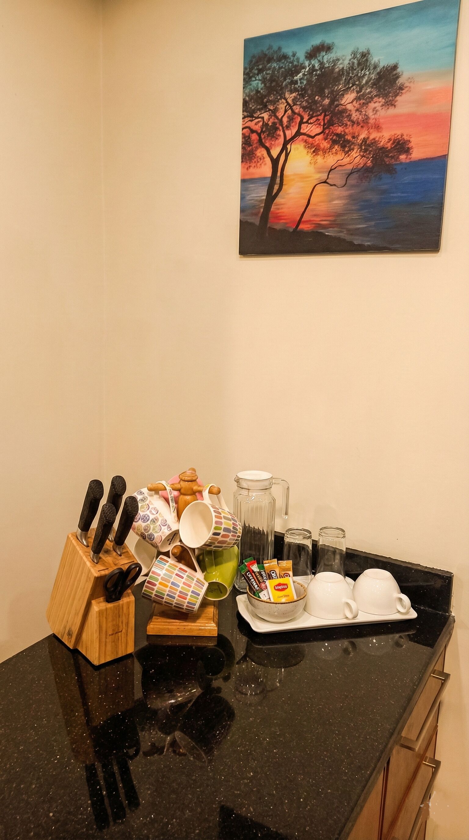 A kitchen counter with a knife block, mugs, a coffee maker, and a painting of a sunset.