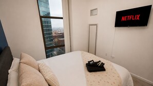 A hotel room with a bed, a TV displaying 'NETFLIX,' a window with a city view, and a door. - Grant Park 2BR Condo with Balcony Skyline View & Gym (Chicago)