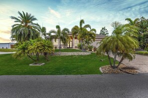 A residential property with a well-maintained lawn, palm trees, and a driveway leading to a house with a tiled roof. - Easy Living Pool House – Relax, Swim, Enjoy (Cape Coral)