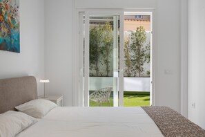 Ein Schlafzimmer mit einem Bett, einem Nachttisch, einer Lampe, einem Sessel und einer Glasgleittür, die auf einen Balkon mit Bepflanzung führt.
