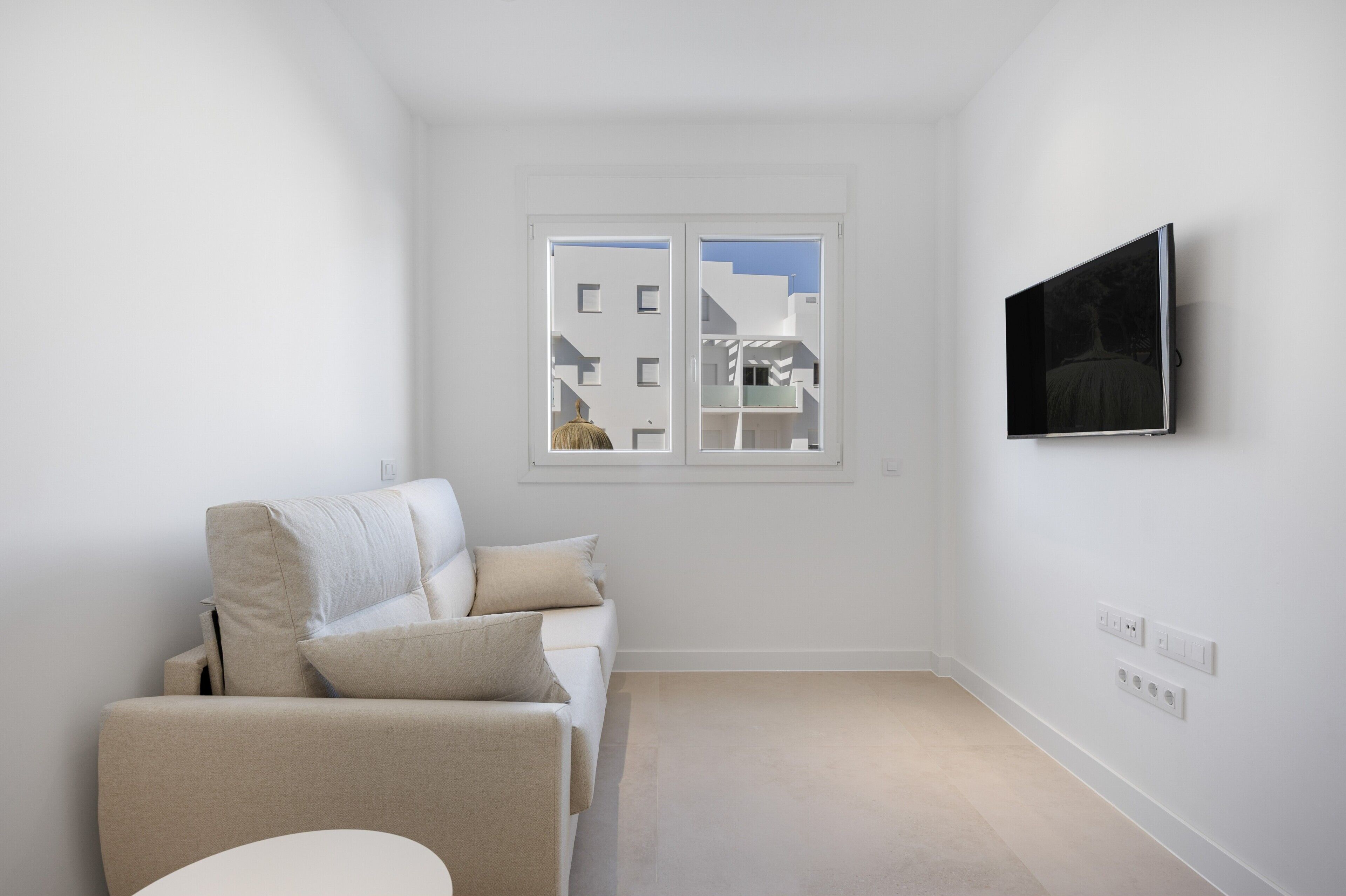Un soggiorno minimalista con un divano, una televisione a parete e una finestra che offre vista sugli edifici.