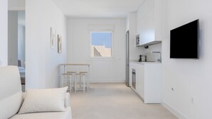 A modern, minimalist living space with a sofa, a small dining area, a kitchen with a TV, and a large window. - Apartment 'Los Drogos, Building A, Penthouse A' with shared pool, garden and Wi-Fi (Chiclana de la Frontera)