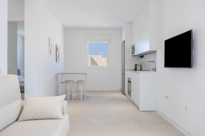Een moderne, minimalistische woonruimte met een bank, een klein eethoekje, een keuken met een televisie en een groot raam.