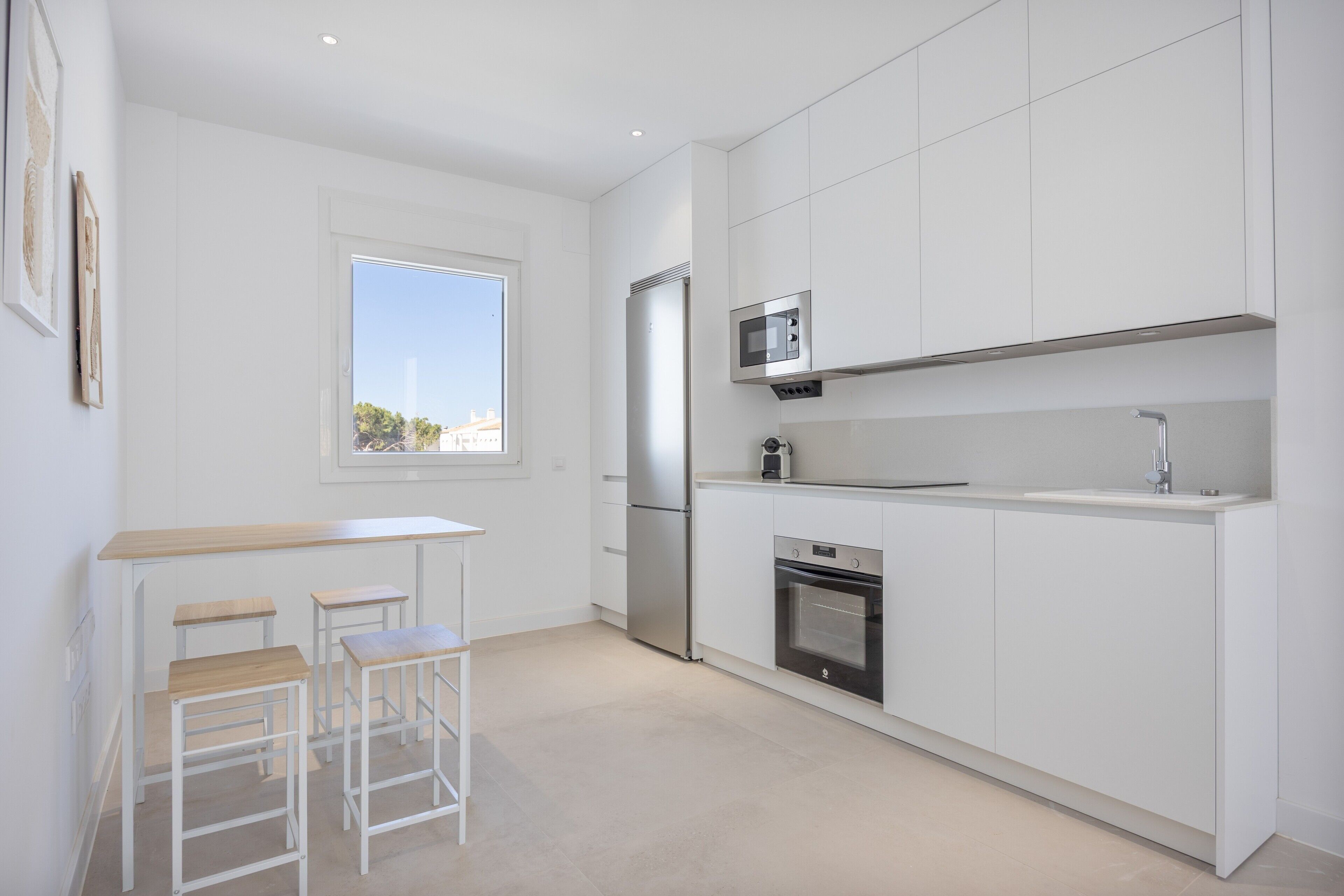 A modern kitchen with white cabinetry, a stainless steel refrigerator, a microwave, and a built-in oven. There are three wooden stools and a window providing a view outside.