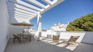 An outdoor patio with a table and chairs, a sun umbrella, and a view of a white building with a tree. - Apartment 'Los Drogos, Building A, Penthouse A' with shared pool, garden and Wi-Fi (Chiclana de la Frontera)