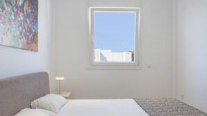 A bedroom with a bed, a nightstand, a lamp, a window, and a painting on the wall. - Apartment 'Los Drogos, Building A, Penthouse A' with shared pool, garden and Wi-Fi (Chiclana de la Frontera)