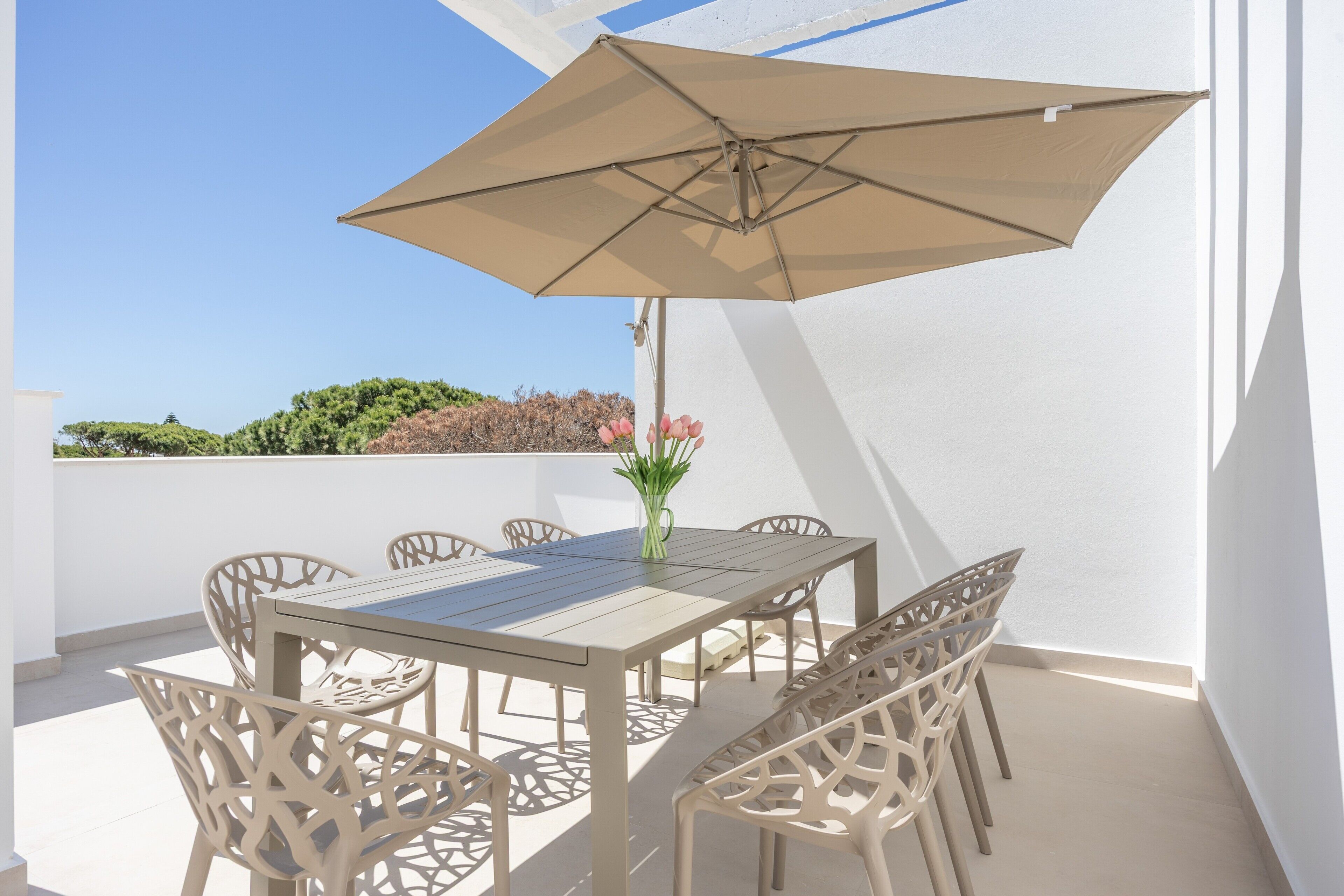Un patio con tavolo e sedie, un ombrellone beige e un vaso con fiori.