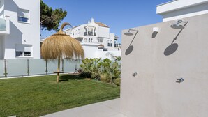 A modern outdoor area with a thatched umbrella, a concrete shower area, and a neatly trimmed lawn. - Apartment 'Los Drogos, Building A, First A' with Shared Pool, Garden and Wi-Fi (Chiclana de la Frontera)