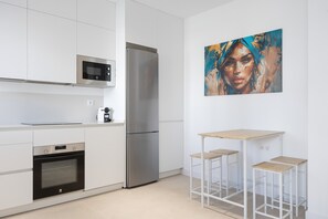 Eine moderne Küche mit weißen Schränken, einem Edelstahlkühlschrank, einer Mikrowelle und einem Backofen. Ein kleiner Holztisch mit zwei Hocker steht daneben.