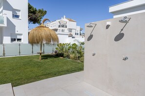 A modern outdoor area with a thatched umbrella, a concrete shower area, and a neatly trimmed lawn. - Apartment 'Los Drogos, Building A, First Floor B' with Pool, Garden & Shared Wi-Fi (Chiclana de la Frontera)