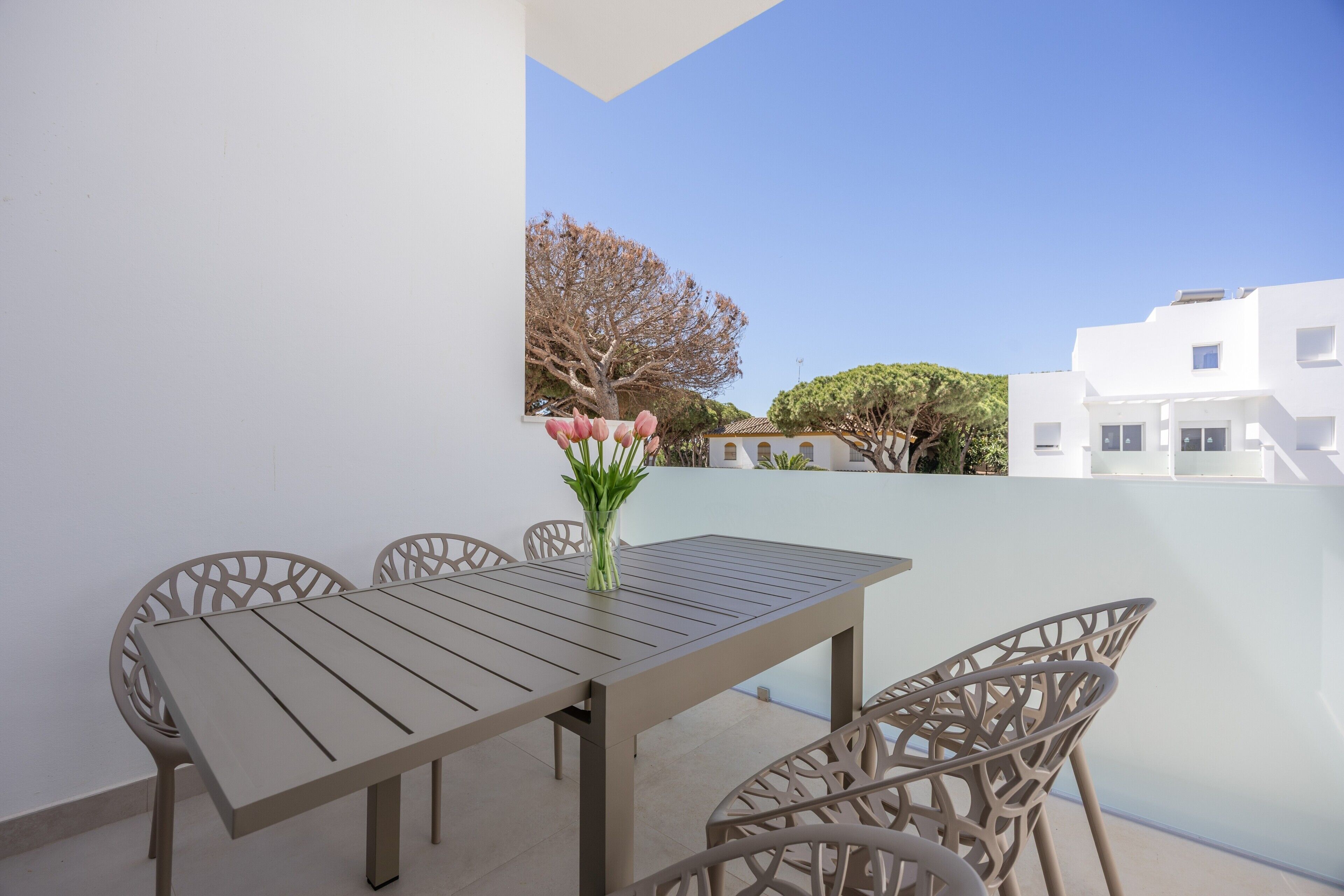 A balcony with a table and chairs, a vase with flowers, and a view of white buildings and trees.