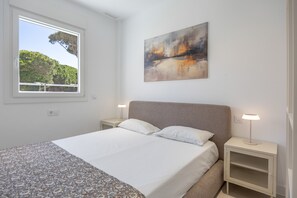 Un dormitori amb llit, taules de nit, finestra amb vista a arbres i un quadre a la paret.
