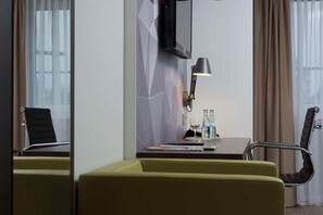 Une chambre d’hôtel équipée d’un bureau, de deux chaises, d’une télévision, d’une lampe et d’une fenêtre avec des rideaux.