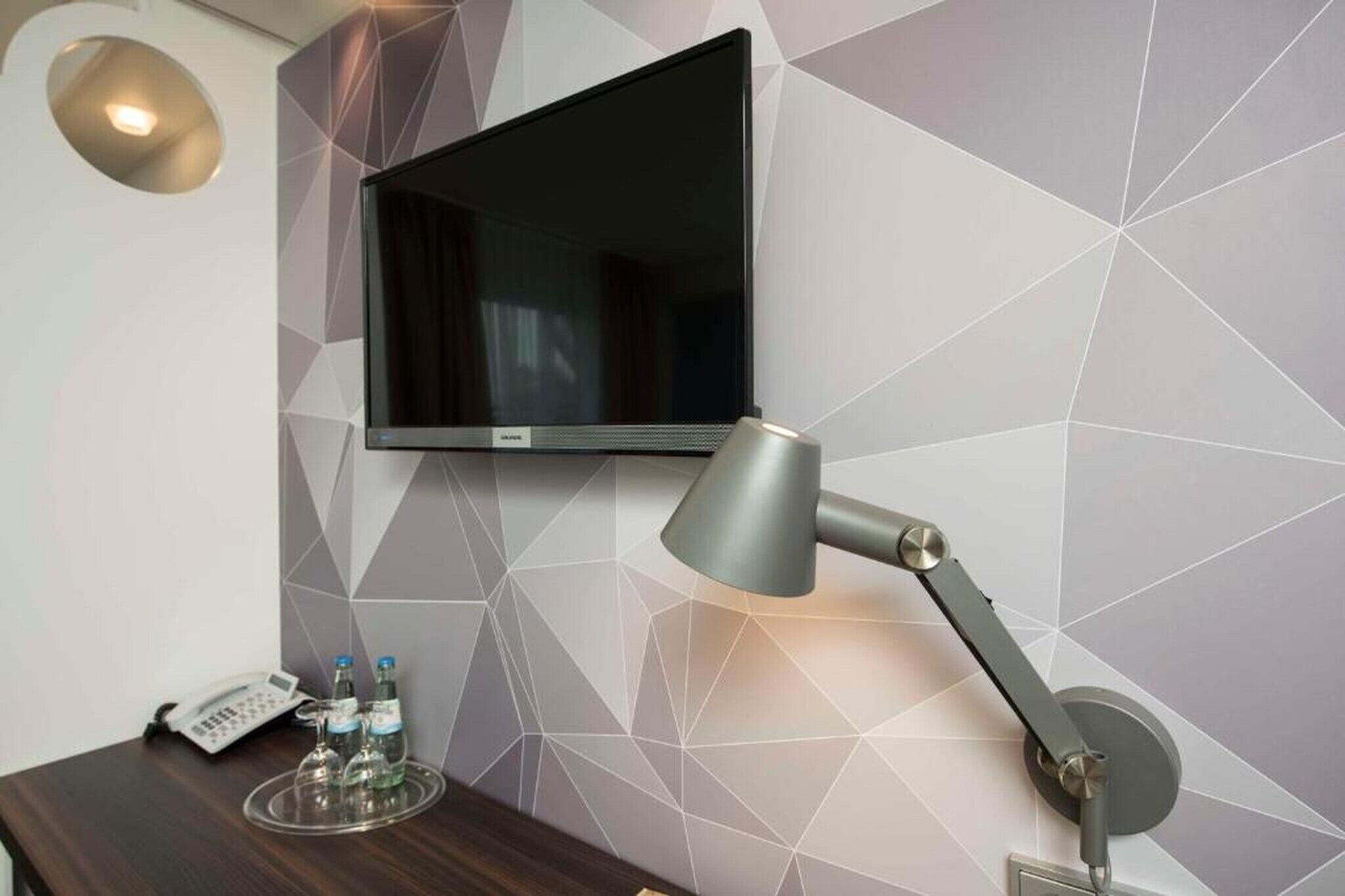 Camera d'albergo con TV a schermo piatto montata a parete, una scrivania con lampada, un telefono e un vassoio con bottiglie.