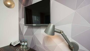 Camera d'albergo con TV a schermo piatto montata a parete, una scrivania con lampada, un telefono e un vassoio con bottiglie.
