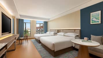 Een hotelkamer met een grote televisie, twee bedden, een bank, een eettafel en uitzicht op gebouwen.