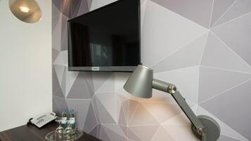 A hotel room with a flat-screen TV mounted on the wall, a desk with a lamp, a telephone, and a tray with bottles.