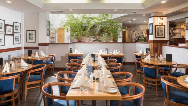 A restaurant with multiple tables and chairs, a central plant display, framed pictures on the walls, and a bar area.