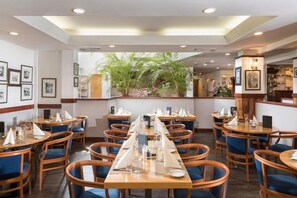 Un restaurant doté de nombreuses tables et chaises, d’un espace central aménagé avec des plantes, de tableaux encadrés aux murs et d’un bar.
