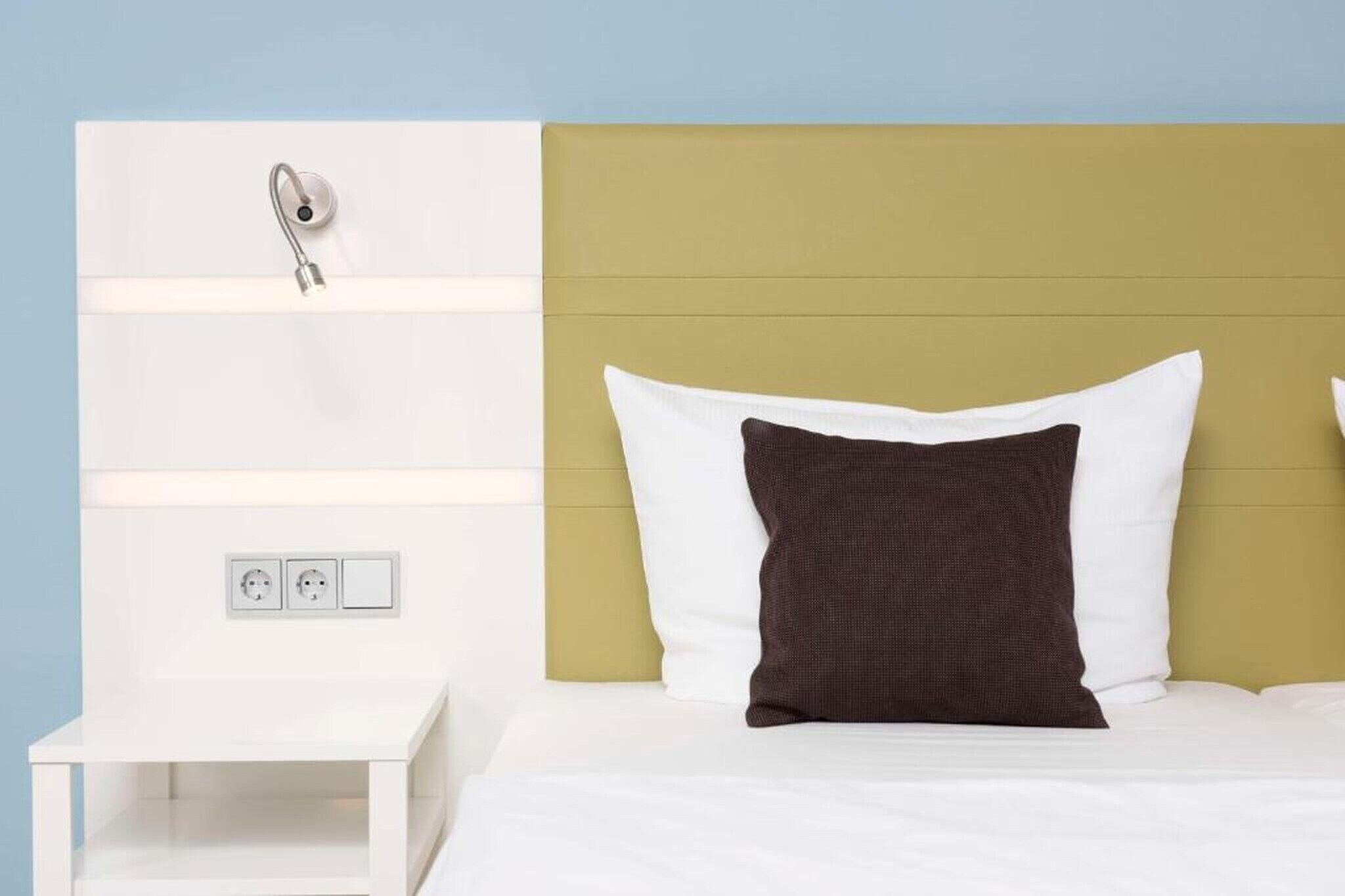 Una cama con almohadas blancas y marrones, una mesita de noche blanca y un aplique de pared.