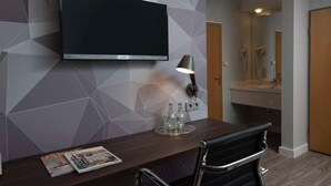 A hotel room with a flat-screen TV mounted on the wall, a desk with a chair, a lamp, and a magazine on the desk.