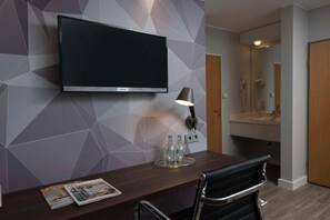 Habitación de hotel con televisor de pantalla plana montado en la pared, escritorio con silla, lámpara y una revista sobre el escritorio.