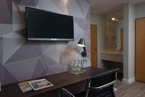 Une chambre d’hôtel équipée d’un téléviseur à écran plat fixé au mur, d’un bureau avec une chaise, d’une lampe et d’un magazine posé sur le bureau.