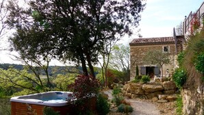 A stone building with a gravel path leading to a hot tub, surrounded by greenery and a view of the countryside.