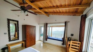 Un dormitorio con cama, techo de madera, un banco, armario, ventana con cortinas y vistas al exterior.